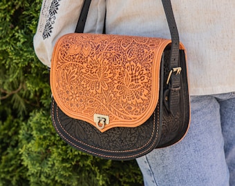 May include: A brown leather satchel with intricate floral patterns embossed on the front flap. The bag has a black leather base and a black adjustable strap.