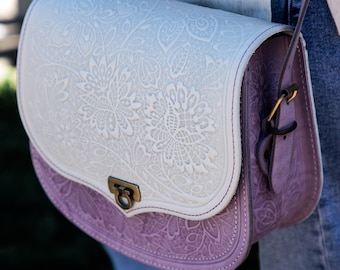 Könnte beinhalten: Eine weiße und lilafarbene Lederschultertasche mit einem floralen Prägemuster. Die Tasche hat einen Klappverschluss mit einer Messing-Schnalle.