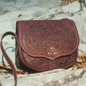 May include: A brown leather satchel bag with an intricate floral and bird design embossed on the front. The bag has a long adjustable strap and a flap closure.