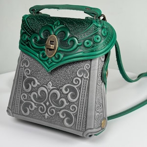 Könnte beinhalten: Eine handgefertigte Ledertasche mit grünem Überschlag und Griff sowie einem grauen Korpus. Der Überschlag weist geprägte florale Designs und einen Messingverschluss auf. Der Taschenkorpus ist mit aufwendigen Schnörkeln verziert.
