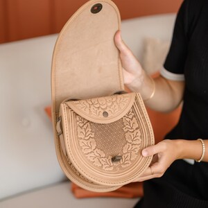 May include: A tan leather saddlebag with a decorative floral pattern and a flap closure. The bag is open, revealing the interior. The bag is held by a person. The bag is a light brown color.
