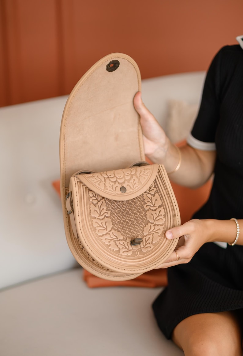 May include: A tan leather saddlebag with a decorative floral pattern and a flap closure. The bag is open, revealing the interior. The bag is held by a person. The bag is a light brown color.