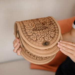 May include: A beige leather saddlebag with a detailed floral and leaf pattern. The bag has a curved shape, a clasp closure, and a layered design. The bag is held by a person.