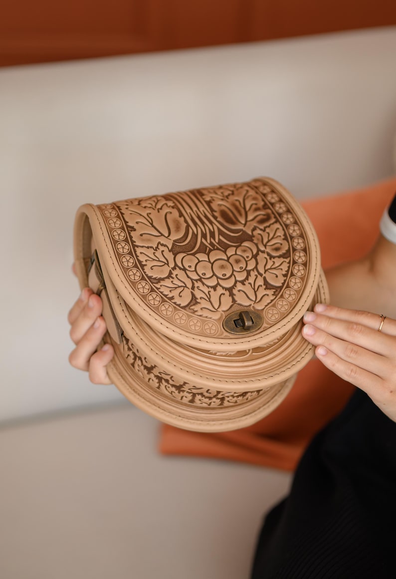 May include: A beige leather saddlebag with a detailed floral and leaf pattern. The bag has a curved shape, a clasp closure, and a layered design. The bag is held by a person.
