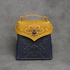 Puede incluir: Un peque&ntilde;o bolso de mano de cuero azul y amarillo con intrincados dise&ntilde;os florales en relieve. El bolso tiene un asa superior y un cierre de lat&oacute;n.