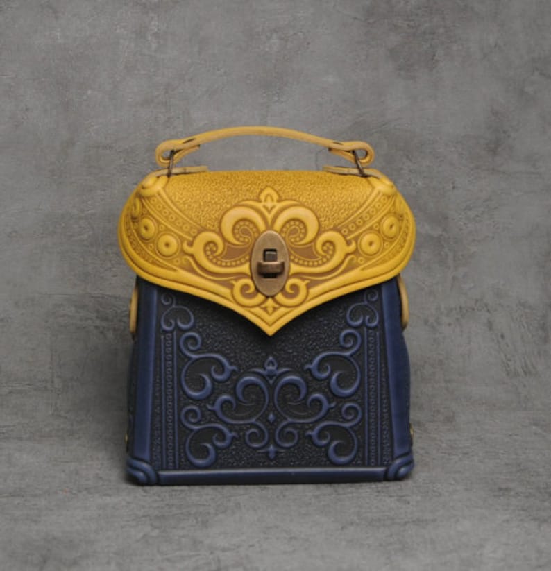 Puede incluir: Un peque&ntilde;o bolso de mano de cuero azul y amarillo con intrincados dise&ntilde;os florales en relieve. El bolso tiene un asa superior y un cierre de lat&oacute;n.