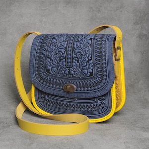 May include: A blue leather crossbody bag with yellow trim and intricate floral embossed detailing. The bag has a flap closure with a metal clasp and a yellow adjustable shoulder strap.