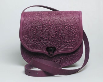 May include: A plum-colored leather crossbody bag with a floral embossed design. The bag features a curved flap, a long adjustable strap, and a metal clasp closure. The bag's dimensions are approximately 10 inches wide, 8 inches high, and 3 inches deep.