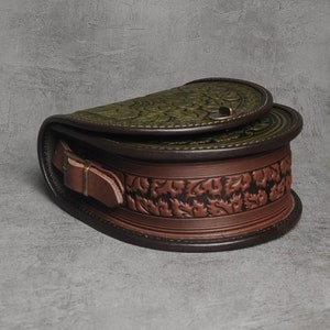 May include: A brown leather satchel bag with a green floral embossed design. The bag has a flap closure with a metal buckle and a brown leather strap.