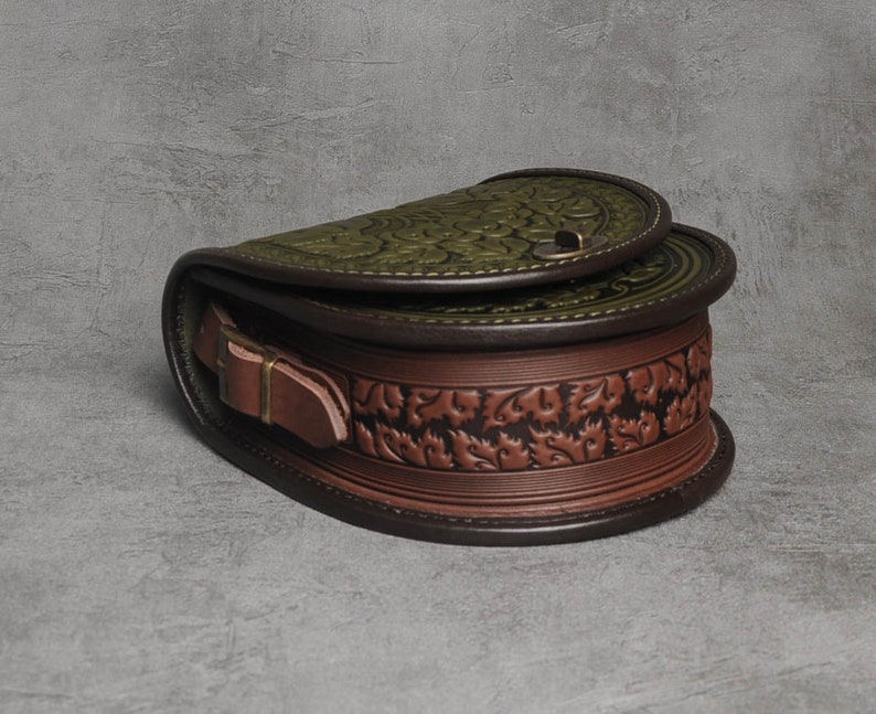 May include: A brown leather satchel bag with a green floral embossed design. The bag has a flap closure with a metal buckle and a brown leather strap.