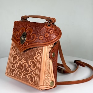 Może przedstawiać: Ręcznie wykonany brązowy skórzany plecak z wytłoczonymi wzorami kwiatowymi. Torba posiada górny uchwyt, regulowane paski i bezpieczne zapięcie na zatrzask. Skóra ma bogaty, ciepły odcień, a design prezentuje szczegółowe rzemiosło.