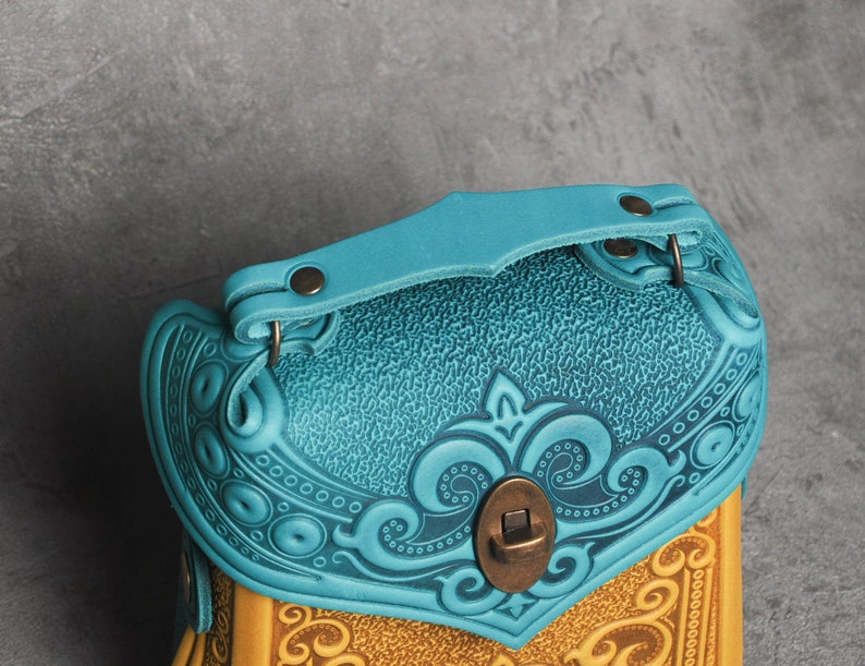 Puede incluir: Un bolso de cuero azul turquesa con intrincados dise&ntilde;os florales en relieve y un cierre de lat&oacute;n. El bolso tiene un asa superior y un cierre de solapa.