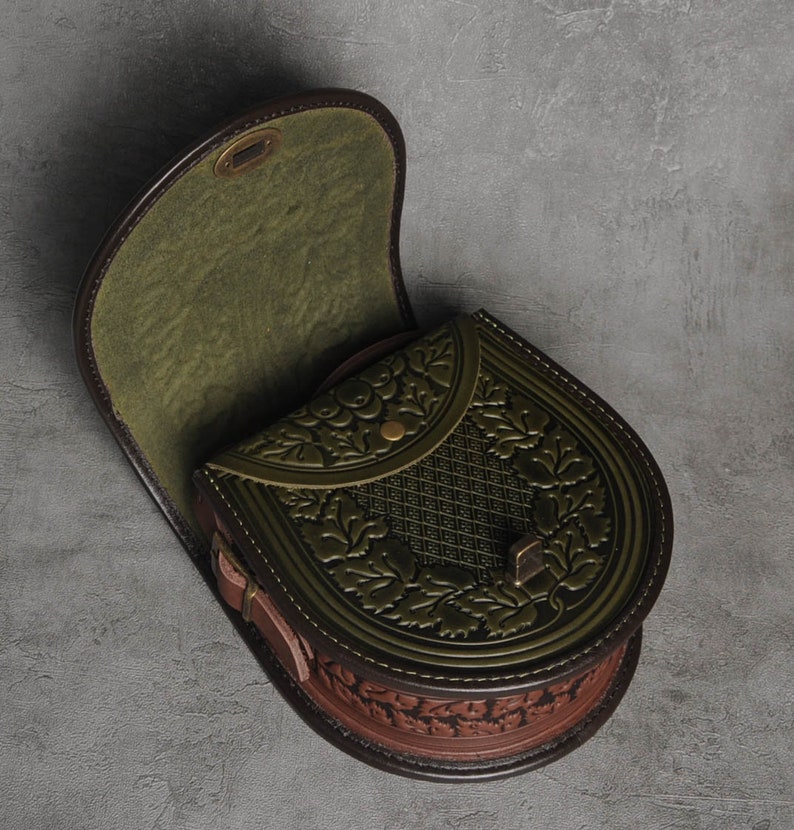 May include: A green leather satchel with a brown leather flap and floral embossed details. The bag has a top handle and a buckle closure.