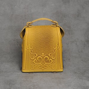 Puede incluir: Una peque&ntilde;a mochila de cuero amarilla con intrincados dise&ntilde;os florales en relieve en la parte delantera. La mochila tiene un asa superior y un cierre de solapa.