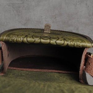 May include: A green leather satchel bag with a flap closure and a brass buckle. The bag has a floral embossed design and a brown leather lining.