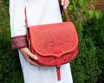 May include: A red leather satchel bag with an intricate floral and bird design. The bag has a long strap and a flap closure.