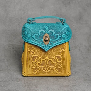 Puede incluir: Un peque&ntilde;o bolso de mano de cuero amarillo con intrincados dise&ntilde;os florales en relieve. La solapa del bolso es azul turquesa con un dise&ntilde;o floral similar y un cierre de lat&oacute;n.