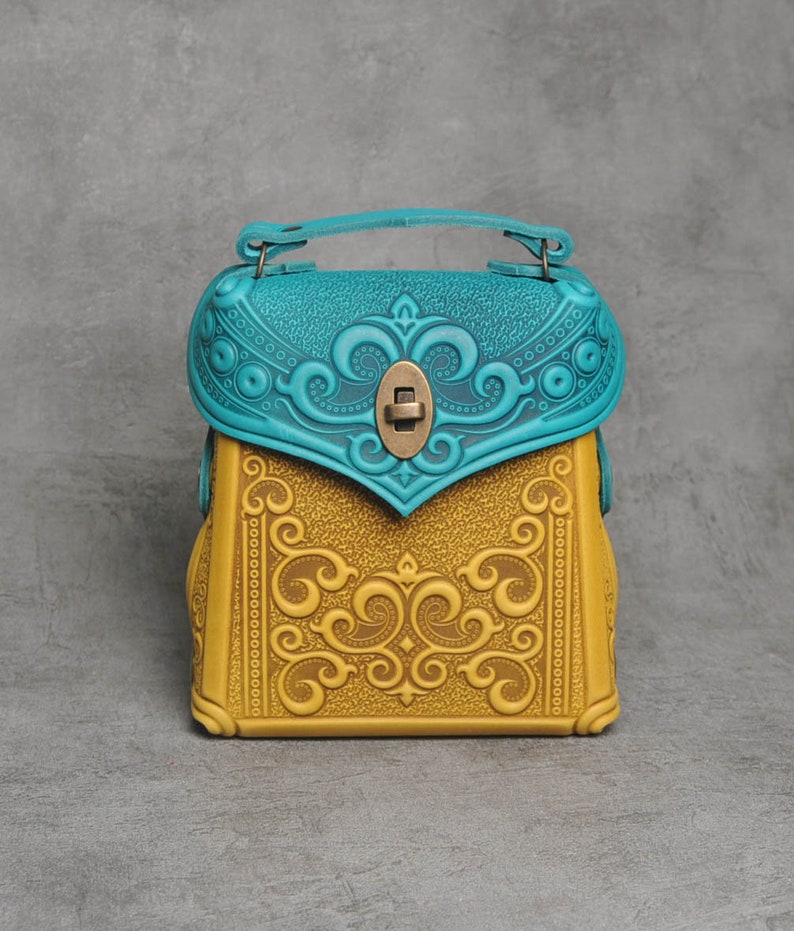 Puede incluir: Un peque&ntilde;o bolso de mano de cuero amarillo con intrincados dise&ntilde;os florales en relieve. La solapa del bolso es azul turquesa con un dise&ntilde;o floral similar y un cierre de lat&oacute;n.