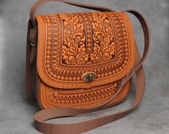 May include: A brown leather satchel bag with intricate floral tooling and a brown leather strap. The bag has a flap closure with a metal buckle.