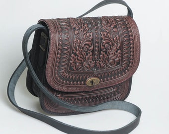 May include: A brown leather crossbody bag with intricate floral embossed detailing. The bag has a flap closure with a metal clasp and a black adjustable strap.