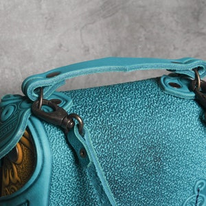 Puede incluir: Un bolso de mano de cuero azul turquesa con asa superior y una correa larga ajustable. El bolso tiene una superficie texturizada y detalles en relieve intrincados.