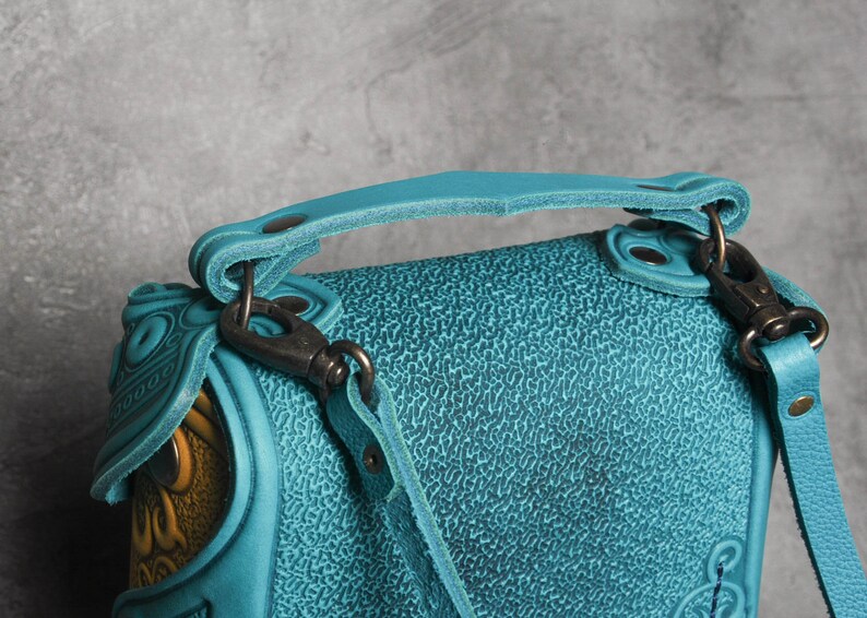 Puede incluir: Un bolso de mano de cuero azul turquesa con asa superior y una correa larga ajustable. El bolso tiene una superficie texturizada y detalles en relieve intrincados.