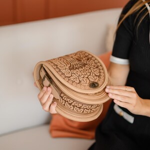 May include: A light brown leather saddlebag with intricate floral and leaf carvings. The bag has a curved shape, a metal clasp, and a layered design. The bag is held open, revealing the interior.