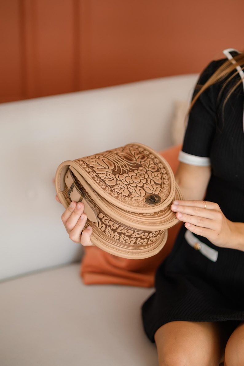 May include: A light brown leather saddlebag with intricate floral and leaf carvings. The bag has a curved shape, a metal clasp, and a layered design. The bag is held open, revealing the interior.