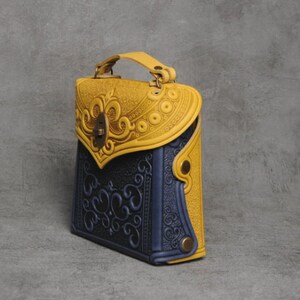 Puede incluir: Un peque&ntilde;o bolso de mano de cuero amarillo y azul con intrincados dise&ntilde;os florales en relieve. El bolso tiene un cierre de solapa con un cierre de metal decorativo y un asa superior.