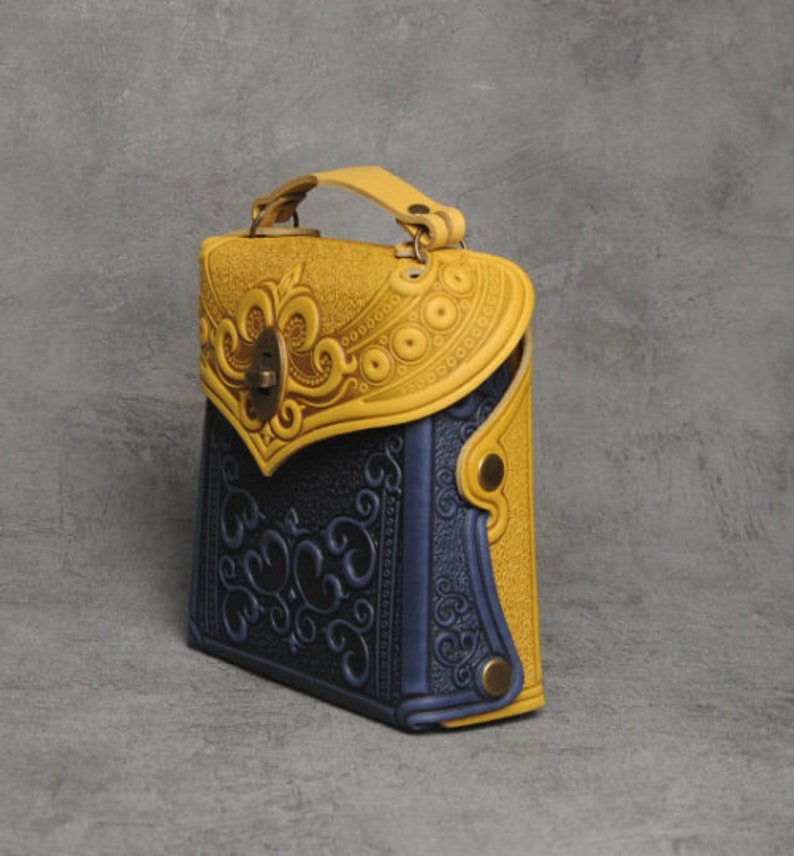 Puede incluir: Un peque&ntilde;o bolso de mano de cuero amarillo y azul con intrincados dise&ntilde;os florales en relieve. El bolso tiene un cierre de solapa con un cierre de metal decorativo y un asa superior.