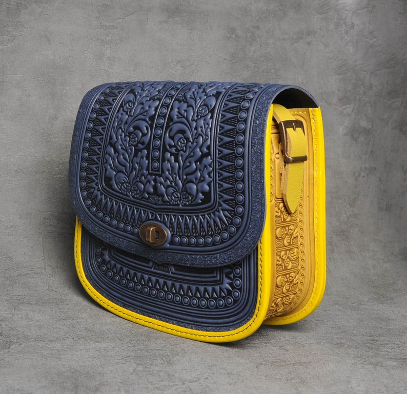 May include: A blue and yellow leather satchel bag with a floral embossed design. The bag has a flap closure with a brass buckle and a yellow strap.