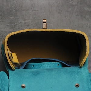 Puede incluir: Un bolso de cuero azul turquesa y amarillo con un cierre de lat&oacute;n. El bolso est&aacute; abierto y el interior es visible.