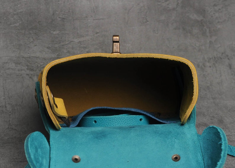 Puede incluir: Un bolso de cuero azul turquesa y amarillo con un cierre de lat&oacute;n. El bolso est&aacute; abierto y el interior es visible.