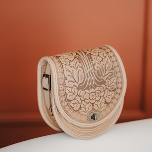 May include: A beige leather saddlebag with a floral embossed design. The bag features a curved shape, a buckle closure, and a decorative border. The bag is made of light-colored leather and has a silver-colored clasp.