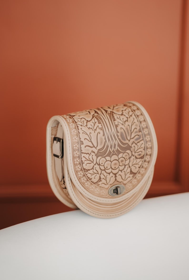 May include: A beige leather saddlebag with a floral embossed design. The bag features a curved shape, a buckle closure, and a decorative border. The bag is made of light-colored leather and has a silver-colored clasp.