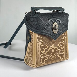Könnte beinhalten: Handgefertigte Leder-Umhängetasche mit geprägtem Design. Die Tasche hat einen schwarzen Deckel mit Griff und einen beigefarbenen Korpus mit kunstvollen Verzierungen. Sie hat einen Metallverschluss und einen langen, verstellbaren Riemen.