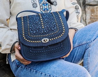 May include: A dark blue leather crossbody bag with intricate embossed floral designs and gold-toned studs. The bag has a flap closure with a gold-toned buckle.