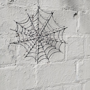 May include: A black spiderweb decoration suspended against a white painted brick wall. The web is constructed from thin, dark lines radiating from a central point, creating a spooky Halloween or gothic aesthetic.