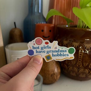 Peut inclure: Un autocollant découpé portant le texte "hot girls have grandma hobbies" en bleu. Le motif comprend des illustrations colorées d'un tambour à broder rose, d'un pelote à épingles rouge, d'aiguilles à tricoter, de pelotes de laine bleue et verte, d'un mètre ruban jaune et d'un motif carré au crochet.