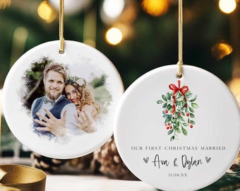 May include: Two round white ceramic Christmas ornaments. One ornament features a photo of a couple, and the other ornament has a watercolor illustration of a sprig of mistletoe with red berries and a red ribbon. The text on the ornament reads "Our First Christmas Married - Ava & Dylan - 11.08.XX".