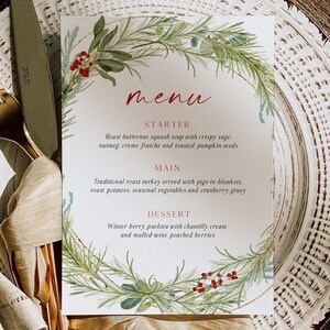 May include: A printed menu card with a watercolor wreath design. The menu features a starter of butternut squash soup, a main course of roast turkey, and a dessert of winter berry pavlova. The card is set on a white plate with gold cutlery.