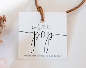 Puede incluir: Etiqueta blanca con texto negro que dice "ready to pop" con un pequeño símbolo de corazón. La etiqueta está atada con una cuerda marrón y tiene el texto "Amber's Baby Shower - August 20, 20XX" en la parte inferior.