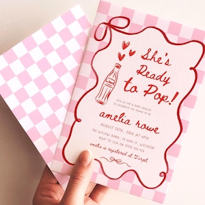 May include: Pink baby shower invitation with a checkered envelope. The invitation features the text "She's Ready to Pop!" in red, along with a soda bottle illustration and event details. The invitation is held in a hand.