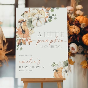 Puede incluir: Un letrero de baby shower sobre un caballete con ilustraciones florales y de calabazas. El letrero dice "A Little Pumpkin Is On The Way!" y da la bienvenida a los invitados al baby shower de Amelia.