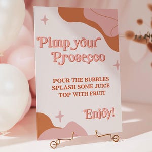 May include: A white sign with pink and brown abstract shapes and gold stars. The sign says "Pimp your Prosecco" and includes instructions to "Pour the bubbles, splash some juice, top with fruit. Enjoy!"