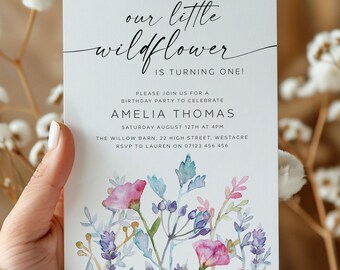 May include: A white invitation card with the text "our little wildflower is turning one!" in elegant script. The card features watercolor floral illustrations in pink, purple, and blue. The invitation is for Amelia Thomas's birthday party.