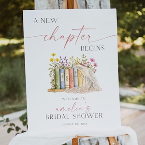 May include: A white sign on an easel with the text "A New Chapter Begins". The sign features a watercolour illustration of books and flowers. The text "Welcome to Amelia's Bridal Shower" is also on the sign.