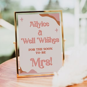 May include: A gold-framed sign with a pink and brown abstract design. The sign reads "Advice & Well Wishes for the soon-to-be Mrs!"