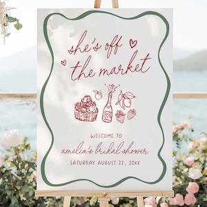 May include: A white sign with a green border and red text that says "She's off the market" with a heart symbol. The sign also has a drawing of a basket of fruit, a bottle of champagne, and strawberries. The text below the drawing says "Welcome to Amelia's Bridal Shower, Saturday August 22, 20XX".