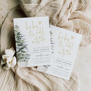 May include: A white baby shower invitation with gold lettering that reads "baby it's cold outside". The invitation features a watercolor illustration of a snowy pine tree. The invitation includes the date, time, and location of the baby shower.